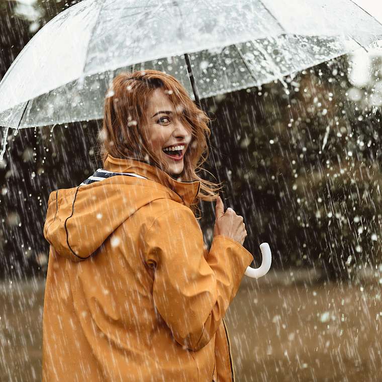 Junge Frau in einer orangenen Jacke und einem Schirm ist ausgelassen fröhlich, obwohl es regnet